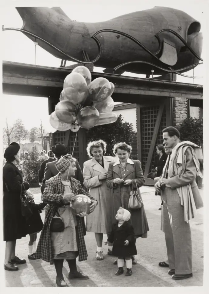 Ernst Hausknost: Luftballonverkäuferin vor dem Gasthaus "Zum Walfisch", um 1955 Eine Frau mit einem Luftballonstrauß zwischen zwei Frauen, einem Mann und einem kleinen Kind, das die Luftballons fasziniert anschaut.